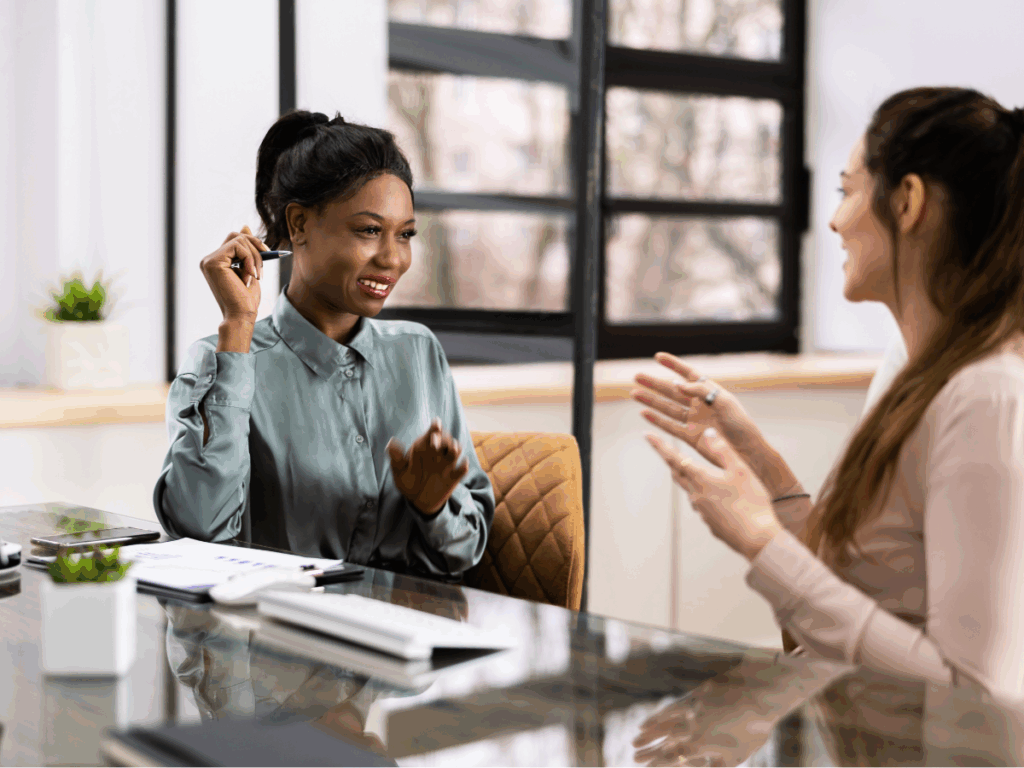 Two women sitting across from each other at a glass desk in a bright office, smiling and talking with documents and a laptop nearby