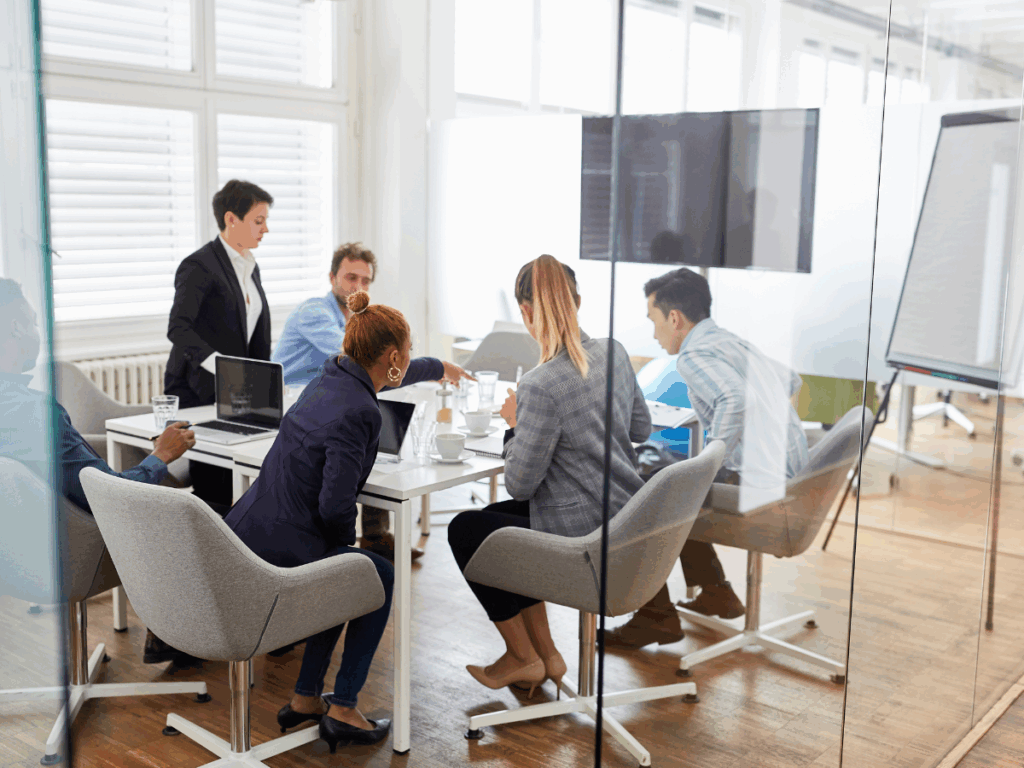 Six professionals in a glass-walled conference room seated around a table with laptops, papers, and glasses during a business meeting